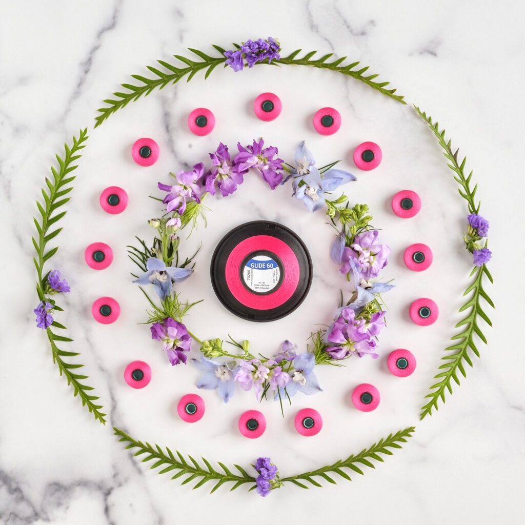 ferns in circle arrangement on marble background surrounding a bright pink spool of thread