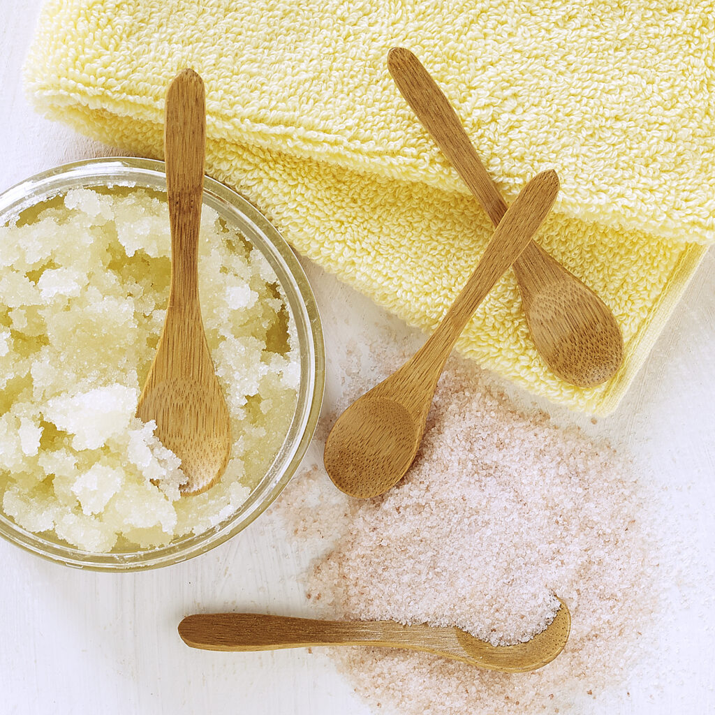 BeeDandy Body sugar scrub with wooden spoon on light neutral background with scattered botanicals