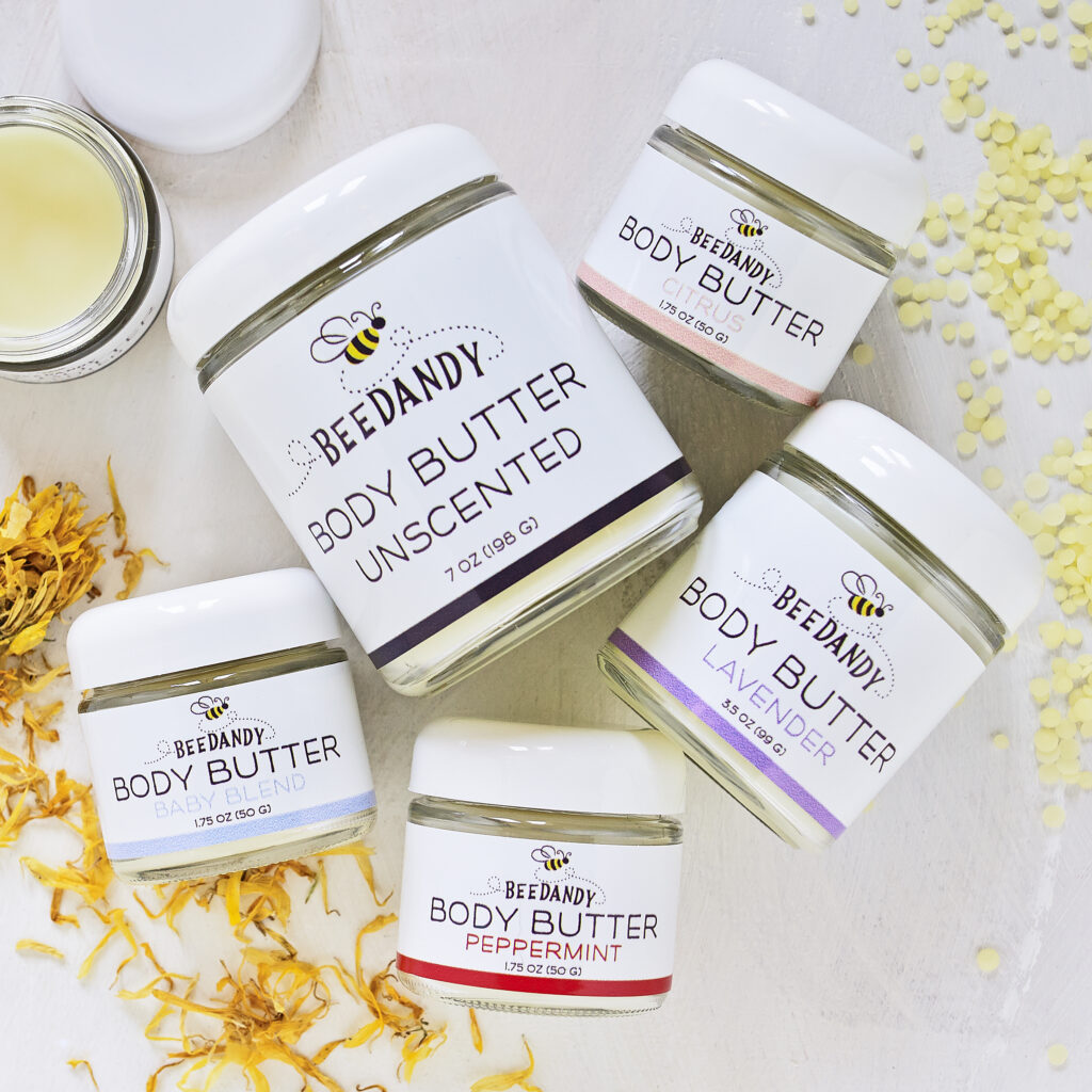 BeeDandy Body Butter jars on light neutral background with scattered botanicals